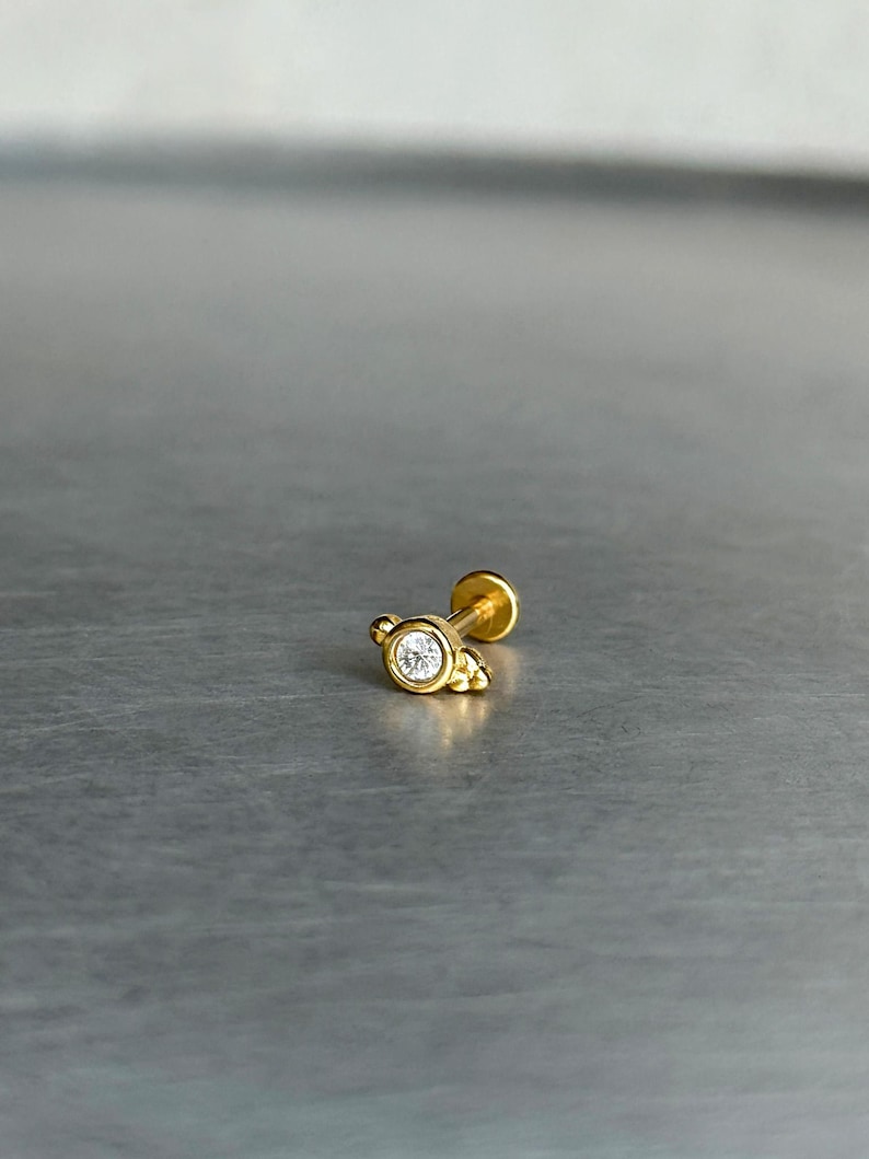 May include: A gold-toned flat back stud earring featuring a central round clear gemstone set in a bezel. The setting includes small decorative gold spheres on the sides and a thin post extending from the back, designed for body piercing jewelry.