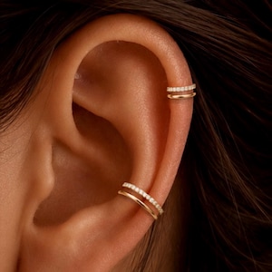 May include: Two gold ear cuffs worn on the helix and conch of an ear. Each cuff features a double-band design, with one smooth polished gold band paired with a second band encrusted with a row of small, clear, sparkling white stones.