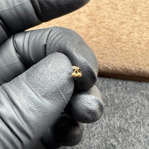 May include: A small, gold-colored cherry stud earring. The earring is held between gloved fingers, with a neutral-toned background. The cherry design features two round, textured cherries and a stem.