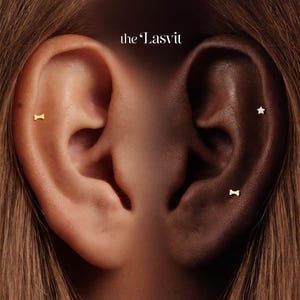 May include: Close-up of two ears, one light and one dark, each adorned with small gold earrings. One ear features a bone-shaped stud, while the other has a star and a bone-shaped stud. The brand name "the Lasvit" is in the top center.