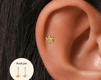 Könnte beinhalten: Ein goldfarbener Ohrstecker in Form einer Sonne mit einem kleinen, hellgrünen Edelstein in der Mitte. Der Stecker befindet sich an einem Ohr. Ein Diagramm zeigt einen Ohrring im Push-Pin-Stil in Gold und Silber.