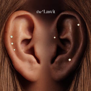May include: Two ears featuring multiple small gold stud earrings with white opal-like stones. The left ear has three studs along the outer cartilage, while the right ear displays two studs. The text "the Lasvit" is centered at the top.