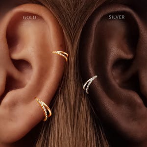 May include: Two ear cuffs are displayed, one gold and one silver. The gold ear cuff is on the left, labeled GOLD, and the silver ear cuff is on the right, labeled SILVER. Each cuff features a double-band design with one band encrusted with clear stones.