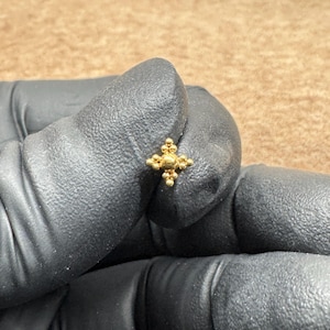 May include: A small, gold-colored stud earring with a cross-shaped design. The earring features a central sphere surrounded by smaller, beaded accents. The earring is held by a gloved hand.
