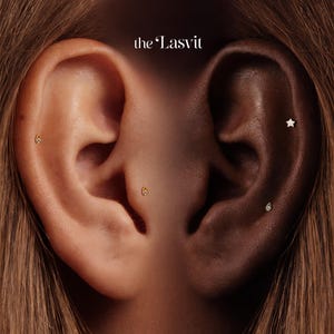 May include: Close-up of two ears, one light-skinned and one dark-skinned, each adorned with delicate earrings. The light-skinned ear features two small gold studs, while the dark-skinned ear showcases a star and a teardrop-shaped earring. The text "the Lasvit" is at the top.