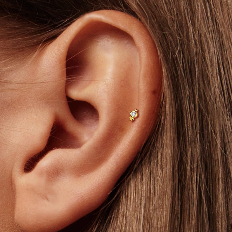 May include: A small gold stud earring featuring a single round clear gemstone set in a circular frame with three tiny gold spheres arranged in a triangular cluster at the base, positioned in the upper cartilage area of an ear.