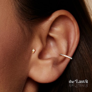 May include: A gold tragus stud featuring a small central clear stone and a gold conch hoop earring set with a row of clear stones. The text "the Lasvit" is visible in the bottom right corner of the image, along with a mirrored reflection.