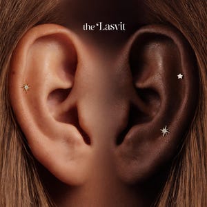 May include: Close-up of two ears, one light-skinned and one dark-skinned, each adorned with delicate earrings. The light ear features a starburst stud, while the dark ear showcases a star and a starburst earring. The brand name "the Lasvit" is visible.