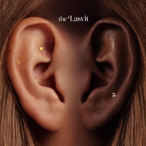 May include: Two ears featuring gold-toned jewelry with the text the Lasvit at the top. The left ear displays a small gold star stud and a gold cherry-shaped stud. The right ear features a single small gold cherry-shaped stud in the lower lobe area.