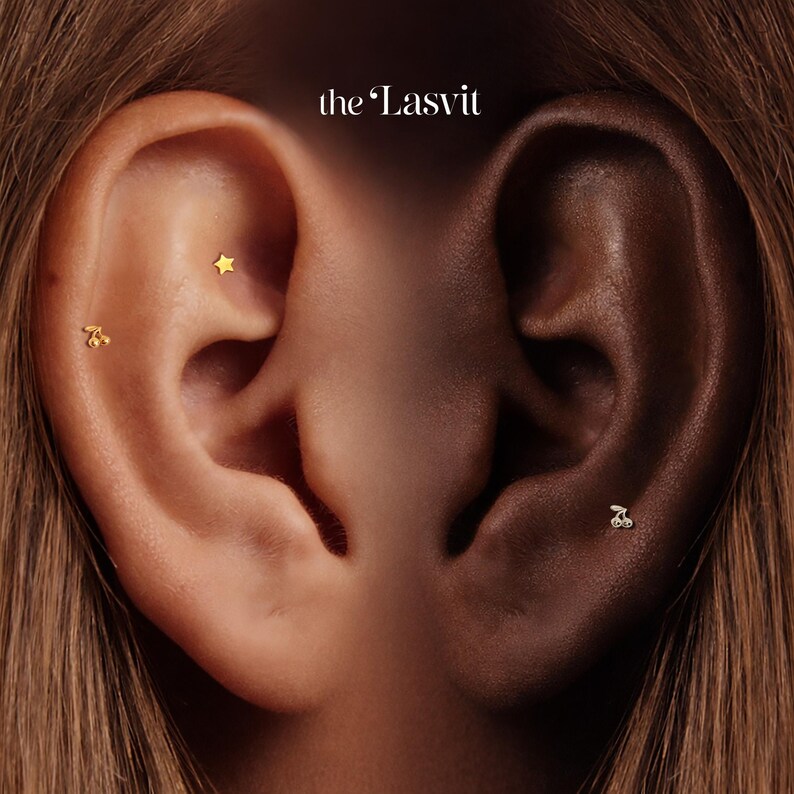 May include: Two ears featuring gold-toned jewelry with the text the Lasvit at the top. The left ear displays a small gold star stud and a gold cherry-shaped stud. The right ear features a single small gold cherry-shaped stud in the lower lobe area.
