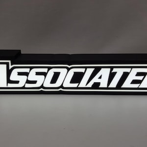 May include: A black sign with white lettering that reads "Team Associated". The sign is illuminated from behind, making the lettering glow.
