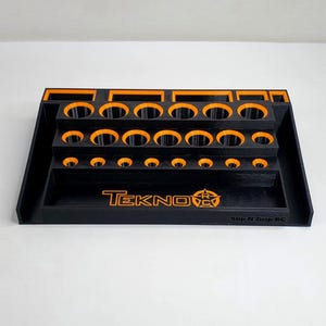 May include: Black and orange Tekno RC tool organizer. The organizer has multiple tiers with various sized circular holes for holding tools. The Tekno logo and "Slip N Grip RC" are printed on the front.
