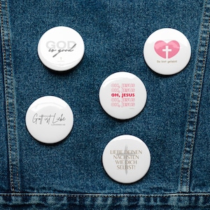 May include: Five white round buttons with religious messages. The buttons have black and gray text. The text on the buttons reads: "God is good", "Gott ist Liebe Johannes 4:8", "Oh, Jesus", "Liebe deinen Nächsten wie dich selbst!" and "Du bist geliebt!" The button with the message "Du bist geliebt!" has a pink heart with a white cross inside.