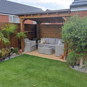 May include: A wooden pergola structure covering a patio area with light gray wicker outdoor furniture, including a sofa, two armchairs, and a matching oval coffee table, set against a brick wall and a wooden fence in a backyard garden.