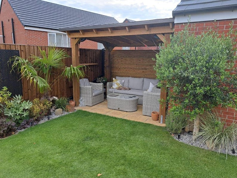 May include: A wooden pergola structure covering a patio area with light gray wicker outdoor furniture, including a sofa, two armchairs, and a matching oval coffee table, set against a brick wall and a wooden fence in a backyard garden.