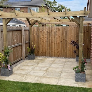 May include: A wooden pergola structure installed over a light-colored square patio area. The pergola features four vertical support posts and horizontal beams. Four dark gray square planters containing green plants are placed at the base.