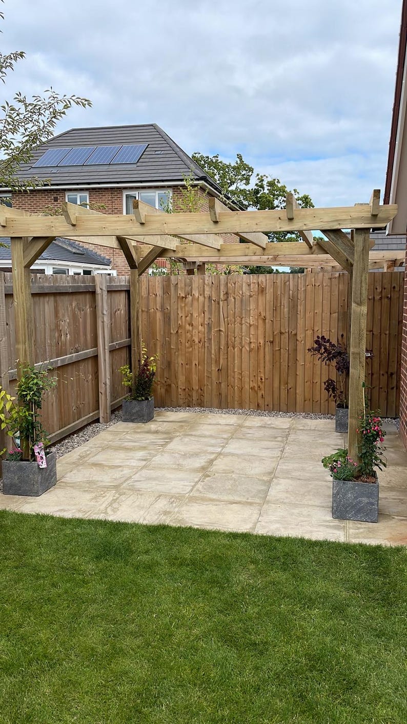 May include: A wooden pergola structure installed over a light-colored square patio area. The pergola features four vertical support posts and horizontal beams. Four dark gray square planters containing green plants are placed at the base.