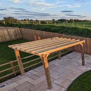 May include: A wooden pergola structure featuring four vertical support posts and a series of horizontal rafters across the top. The frame is constructed from light brown, pressure-treated timber beams, creating an open-roof outdoor garden feature.