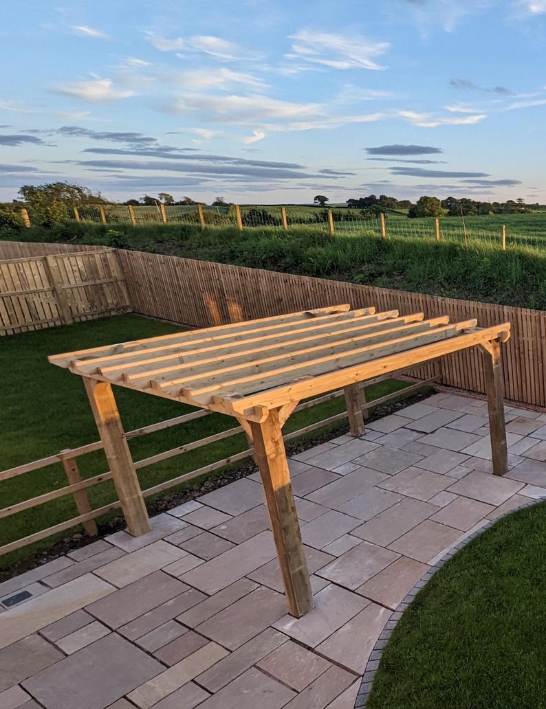 May include: A wooden pergola structure featuring four vertical support posts and a series of horizontal rafters across the top. The frame is constructed from light brown, pressure-treated timber beams, creating an open-roof outdoor garden feature.