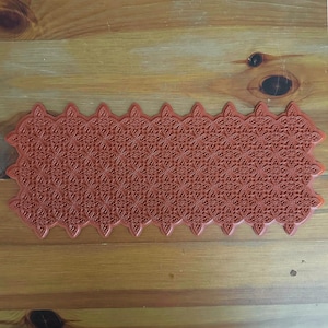 May include: A rectangular, terracotta-colored rubber stamp with a detailed, repeating floral pattern. The stamp has a scalloped edge, and the intricate design is visible across the entire surface. The stamp is designed for crafting and decorative purposes.