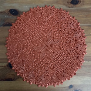 May include: A round, terracotta-colored placemat with a detailed floral and leaf pattern. The placemat has a scalloped edge and is made of a flexible material. The design is symmetrical, with a central floral motif radiating outwards.