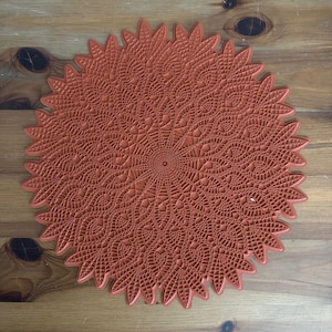 May include: A large, round, terracotta-colored lace doily with a detailed, intricate design. The doily has a raised, textured surface and a scalloped edge.