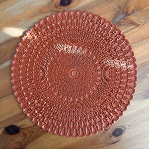 May include: A round, terracotta-colored placemat with a detailed, intricate design of overlapping circles and teardrop shapes.