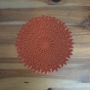 May include: A terracotta-colored silicone trivet with a detailed, intricate lace pattern. The trivet is round and has a raised edge.