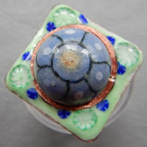 May include: A square ceramic tile with a raised, blue and white floral design in the center. The tile is surrounded by a copper border and a green and white floral design.