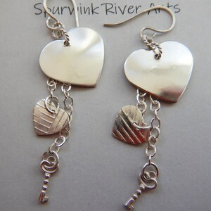 May include: Silver heart earrings with a smaller heart charm and a key charm dangling from each earring. The earrings are labeled "2019  Sourvink River Arts".