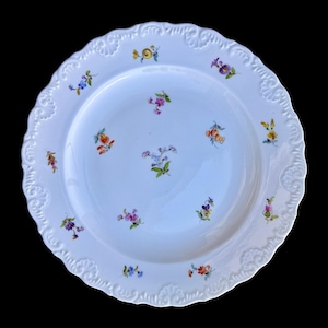 Könnte beinhalten: Weißer Porzellanteller mit gewelltem Rand und geprägtem Blumendesign. Der Teller ist mit kleinen, farbenfrohen, handgemalten Blumen in Lila, Gelb, Orange und Blau verziert. Der Teller ist vor einem schwarzen Hintergrund platziert.