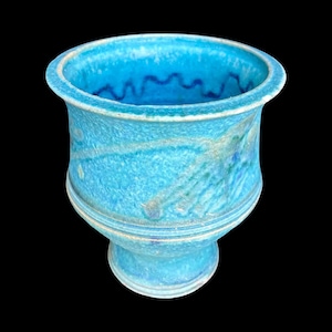 May include: A turquoise ceramic vase with a pedestal base and a wide, fluted rim. The vase has a textured surface with streaks of lighter blue and green. A wavy, dark blue line decorates the inside rim. The vase is set against a black background.