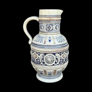 May include: A white and blue ceramic jug with a handle and spout. The jug features intricate embossed designs, including geometric patterns, floral motifs, and a lion's head. The colour scheme includes white, blue, and brown accents.
