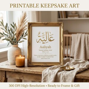 A framed gold and white printable keepsake art print featuring the Arabic name Aaliyah stands on a wooden table next to a vase of dried stems and a lit candle.