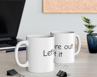 May include: Two white ceramic mugs with black text that reads "Let's it ire out". The mugs are on a light-colored wooden surface, with a laptop and office supplies in the background.
