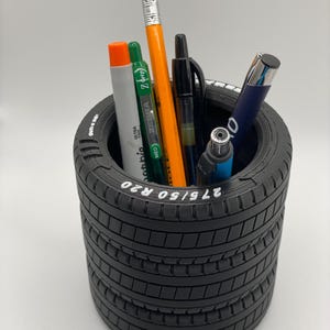 May include: Black tire-shaped desk organizer with a white label that reads "275/50 R20". The organizer is filled with various writing utensils, including pens, pencils, and a lighter.