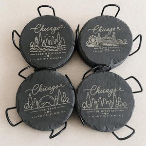 May include: Four round, dark gray slate coasters with black metal handles. Each coaster features a different Chicago landmark design in white, including the skyline, Navy Pier, and The Bean, with the city's name in script.