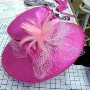 May include: A pink satin hat with a wide brim and a pink feather trim. The hat also features a pink tulle bow.