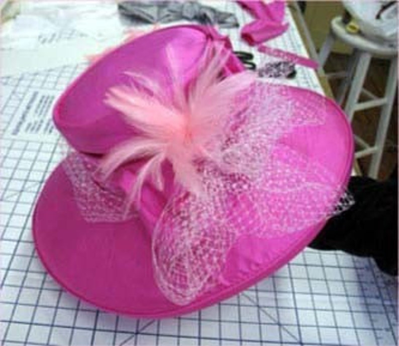 May include: A pink satin hat with a wide brim and a pink feather trim. The hat also features a pink tulle bow.
