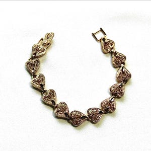 May include: A gold-toned metal bracelet composed of a series of interconnected heart-shaped links. Each heart features an intricate, openwork filigree pattern. The bracelet includes a rectangular clasp mechanism at one end for secure fastening.