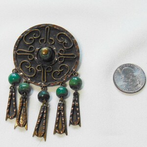 May include: Vintage bronze-toned brooch with a circular design and green bead accents. The brooch features intricate scrollwork and dangling pendants. A quarter coin is next to the brooch for size comparison.