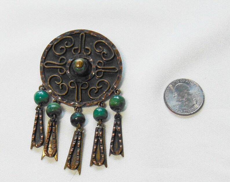 May include: Vintage bronze-toned brooch with a circular design and green bead accents. The brooch features intricate scrollwork and dangling pendants. A quarter coin is next to the brooch for size comparison.