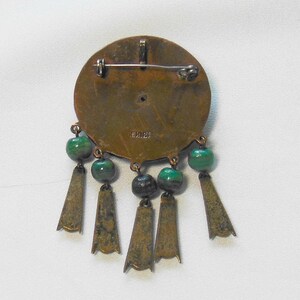 May include: Vintage bronze-toned brooch with a circular design, adorned with green beads and dangling bronze-colored pendants. The brooch has a pin closure on the back and the word "MEXICO" is stamped on the back.