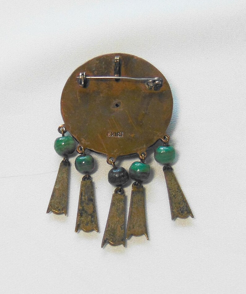 May include: Vintage bronze-toned brooch with a circular design, adorned with green beads and dangling bronze-colored pendants. The brooch has a pin closure on the back and the word "MEXICO" is stamped on the back.