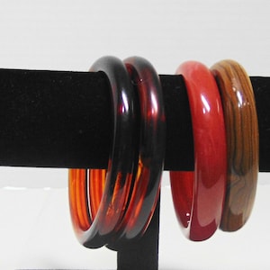 May include: Four colorful bangles displayed on a black velvet jewelry holder. The bangles are in shades of red, brown, and orange. The bracelets are made of a glossy material and are round in shape. The jewelry holder is rectangular.