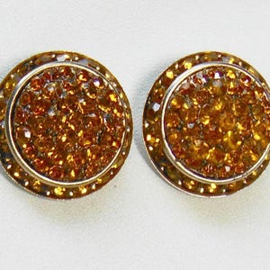 May include: A pair of circular stud earrings featuring a central cluster of amber-colored crystals surrounded by a smooth metallic border. The surface of each earring is densely packed with small, faceted stones that reflect light, creating a textured appearance.