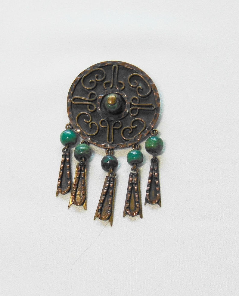 May include: A round, copper-toned brooch with an ornate, embossed design. The brooch features green beaded accents and dangling copper-colored pendants. The central design has a raised, circular element. The overall style is vintage.