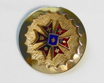 May include: A circular gold-tone metal brooch featuring a textured sunburst border and a central eight-pointed cross. The cross is decorated with red enamel, small gold symbols, and a central blue shield displaying a gold fleur-de-lis emblem.