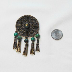 May include: A vintage copper-toned brooch with a circular design and intricate patterns. The brooch features green beads and dangling decorative elements. A quarter is next to the brooch for size comparison.