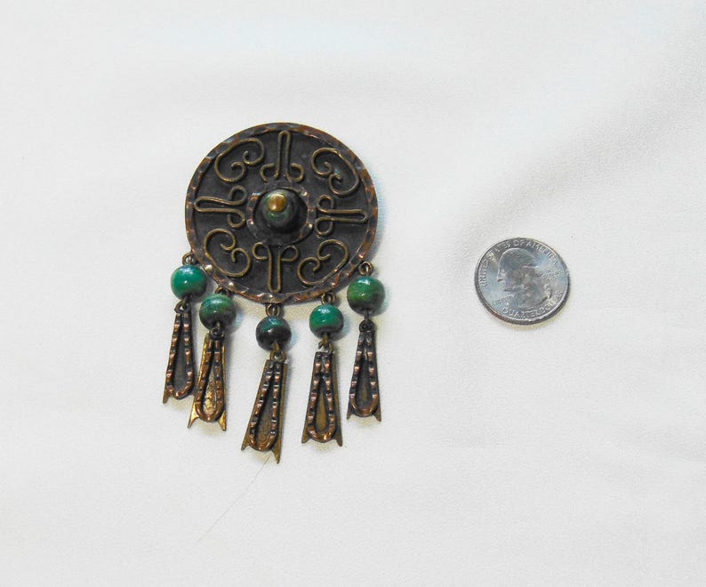 May include: A vintage copper-toned brooch with a circular design and intricate patterns. The brooch features green beads and dangling decorative elements. A quarter is next to the brooch for size comparison.
